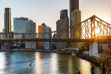 Yacht passing under Brisbane Story Bridge as the sunsets behind the cityscape - obrazy, fototapety, plakaty