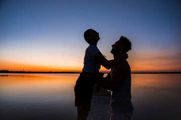 Fototapeta premium Silhouettes of young beautiful couple resting rejoicing at sunrise near lake.