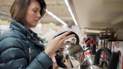 Beautiful brunette woman in coat chooses a kettle in the shop of home appliances