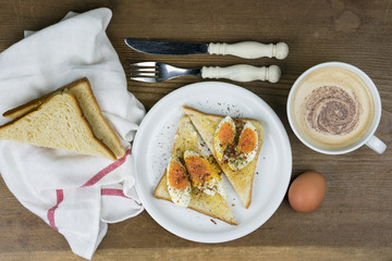 Egg sandwich seasoned with pepper with frothy cappuccino coffee, a knife and fork and sliced bread on a rustic wooden table, overhead view