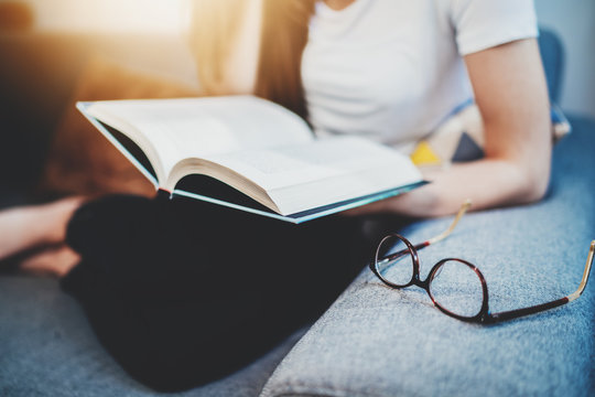 Closeup of young hipster girl reading a book, free time and education concept, woman holding new bestseller book while sitting at home interior on cozy sofa, eyeglasses on sofa