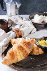 Breakfast with two croissant, butter, cup of coffee, cottage cheese and sliced mango fruit, served on serving wood board with textile napkin on white and gray concrete texture background. Close up.