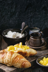 Breakfast with two croissant, butter, cottage cheese, coffee and sliced mango fruit, served on wood chopping board over old wooden background. Close up.