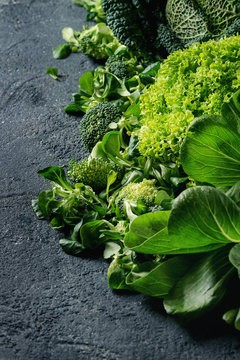Variety of raw green vegetables salads, lettuce, bok choy, corn, broccoli, savoy cabbage as frame over black stone texture background. Space for text