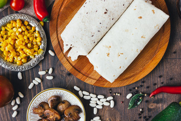 Vegetarian burrito on wooden board over black table surrounded by ingredients.