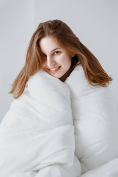 Portrait of smiling beautiful young Caucasian girl woman with long  red hair sitting in bed wrapped covered with  blanket on white background, looking in camera, early morning