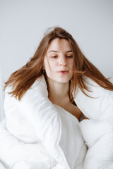 Portrait of beautiful young Caucasian girl woman sitting in bed wrapped covered with blanket on white background, blowing her hair on early morning, lifestyle