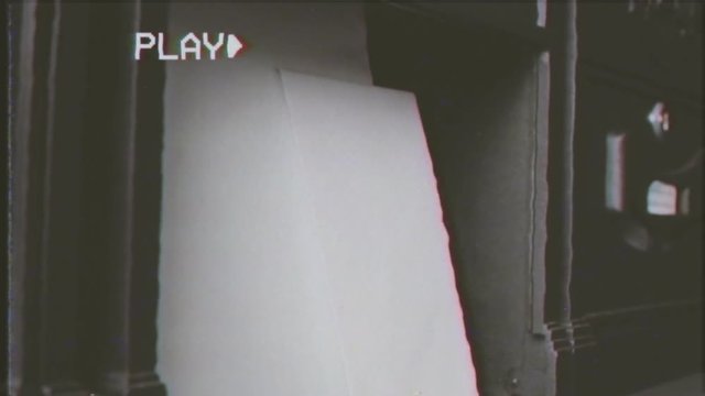 Fake VHS tape: emptying a mail slot (one in a line of wall-mounted mailboxes) full of letters. Indoor close-up shot.

