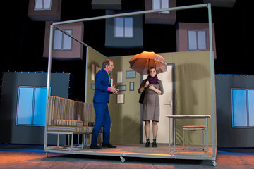 Two actors, brunette woman in a brown dress and a man in a pink shirt and blue suit, playing the role on stage © Kozlik_mozlik