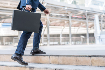 Close up shot of businessman holding leather briefcase while walking upward on the stair outdoor in city. - obrazy, fototapety, plakaty