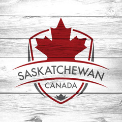 A Canadian province crest on a background of distressed barnboard. The shield features a maple leaf and the main text says Saskatchewan, Canada.