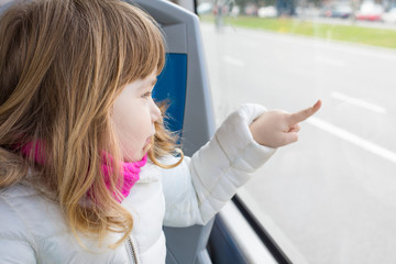 Three years old blonde child sitting in public bus with white coat and pink scarf, touching window...