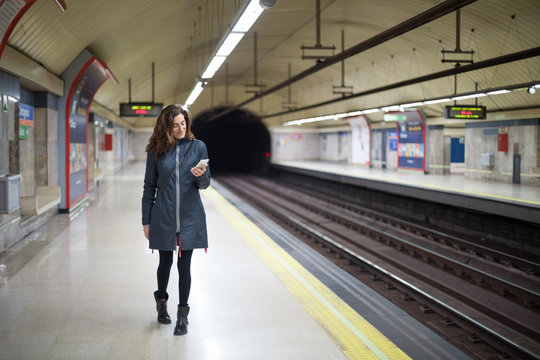 smiling woman with blue coat, walking in platform of subway or underground station of Madrid city, using and typing mobile smart phone
