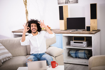 Hipster freelance man showing okay signs to camera while sitting on sofa or couch and working on laptop computer at home alone.