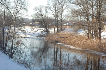 Suspension bridge over a small river against the background of reeds and snow-covered shores.