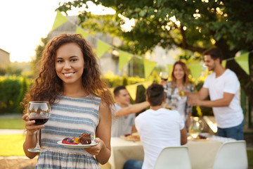 Pretty young woman holding glass of red wine and plate with grilled meat and vegetables