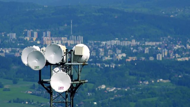 White parabolic antennas on the top of a radio tower, a town and a forest in the blurry background