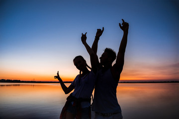 Fototapeta premium Silhouettes of young beautiful couple resting rejoicing at sunrise near lake.
