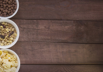 Bowls of corn flakes , chocolate balls and muesli on wooden table