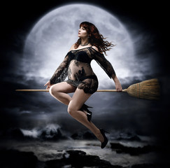 Fashionable redhead pregnant woman flying on a broom in black, transparent and lace peignoir