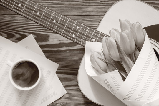 Beautiful spring background music. Guitar, yellow tulips, cup of coffee, musical page on a dark wooden background. Coloring and processing photo with old monochrome style.