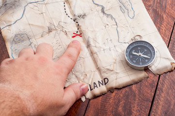 Man point by finger to red cross where is Pirate treasure chest on Fake map with compass on wooden table