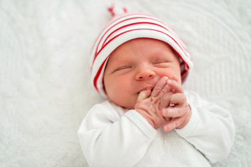 Sleeping newborn baby on back on white bed in a striped white and red cap sucks a finger . Close up