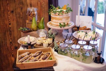 sweet bar with eclairs, lemonade,sweets and white cake  decorated with greenery in area of wedding party