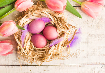 Easter eggs in nest, fresh pink tulips on white wooden background.