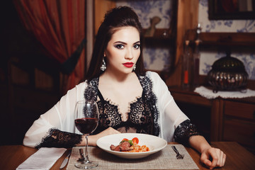 A beautiful girl in her underwear eats spaghetti pasta with meat and wine in a cafe-restaurant. She eats her hands. Dark background. Fashion. Portrait. Close-up.