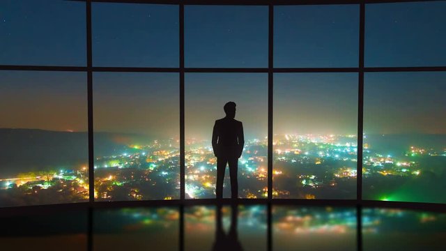 The man stand near the panoramic window on the night city background. Time lapse