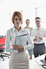 Portrait of confident businesswoman holding book with colleagues in background at office