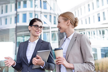 Obraz premium Happy businesswomen with tablet PC and disposable cup conversing outside office building