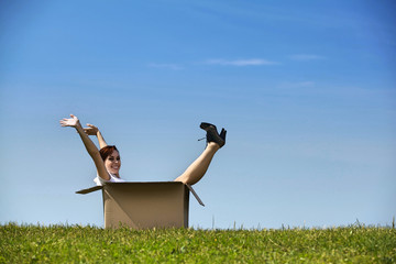 Cheerful young woman sitting in cardboard box at park with arms 