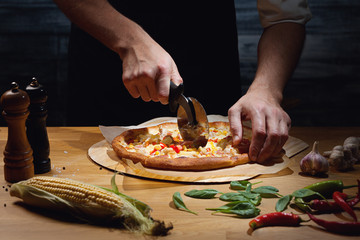 Chef cutting freshly baked hawaiian pizza. Low key shot, close up of hands, some ingredients around on table.