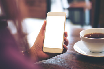Mockup image of hands holding white mobile phone with blank white screen and hot coffee cup in loft cafe