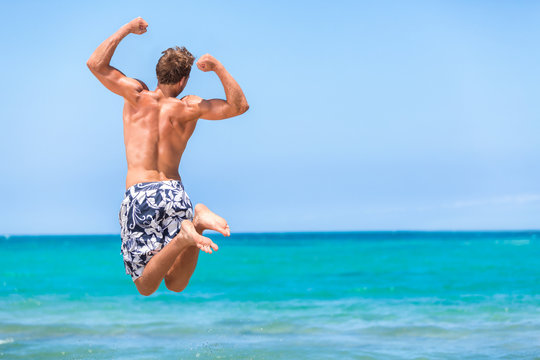 Happy fitness man jumping of goal achievement success on beach. Male athlete topless in swim shorts doing joy jump on ocean background