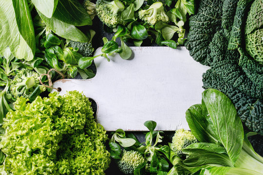 Variety of raw green vegetables salads, lettuce, bok choy, corn, broccoli, savoy cabbage as frame round empty white chopping board. Food background. Top view, space for text
