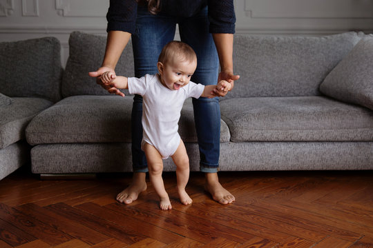 Smiling baby learning to walk in the living room with his mother holding his hands
