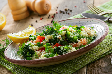 Plate with couscous and vegetables on a rustic wooden table. Traditional eastern healthy meal made of couscous, broccoli, tomato, pepper, onion and dill. Close-up shot.