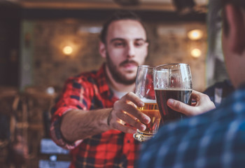 Two bearded hipsters clinking glasses of beer at bar, focus on glasses. Cheers.