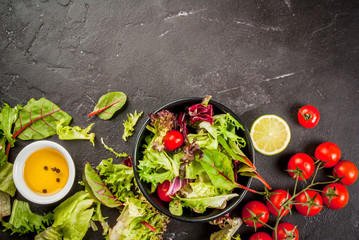 Light salad of green leaves and tomatoes, oil and lime juice. On a black concrete table, with ingredients, copy space top view