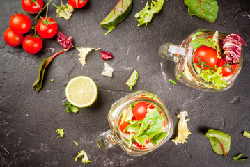 Light dietary healthy lunch: a spring salad in jars, salad leaves, tomatoes, dressed with lime juice and olive oil. On a black concrete table, with ingredients. Two portions. Copy space