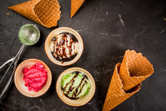 Selection of colorful homemade ice cream: lemon (pistachio) green, berry pink, white vanilla. With a spoon for serving balls, waffle cones, on a slate board, on a black table. Top view, copy space