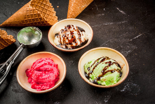 Selection of colorful homemade ice cream: lemon (pistachio) green, berry pink, white vanilla. With a spoon for serving balls, waffle cones, on a slate board, on a black table. Top view, copy space