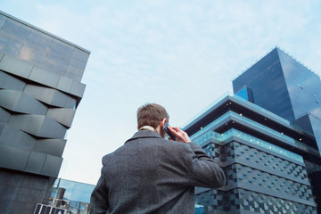 A pretty dressed man talking on a mobile phone in front of an office skyscraper. View from the back from the bottom up.