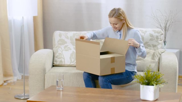 Beautiful Young Woman at Home Opens Cardboard Box while Sitting on a Couch in Her Bright Living Room. She Smiles.  Shot on RED EPIC-W 8K Helium Cinema Camera.