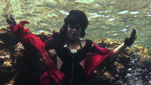 Young girl model free diver underwater in red costume of pirate in Red Sea. Filming a movie. Actress smiling at camera. Extreme sport in marine landscape, coral reefs, ocean inhabitants.