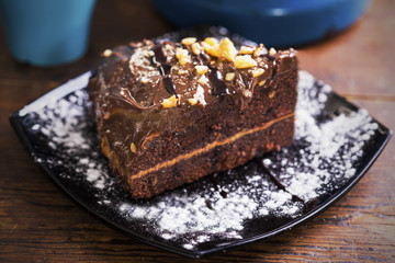 Piece of chocolate cake with icing sugar and nuts served on black plate