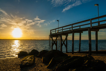 Fototapeta premium The setting sun with clouds in the background with a rocky beach, large algae-covered stones, and a wooden pier in the foreground. West Neck Beach in Huntington, NY, USA.